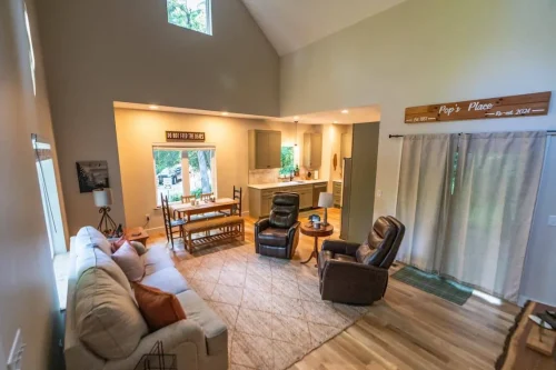 Cozy open-concept living area with vaulted ceiling, sofa, recliners, dining table, and kitchen in the background.