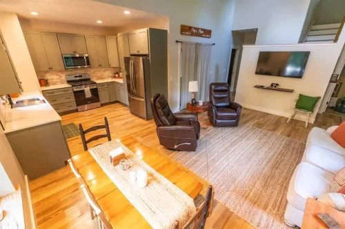 Bright kitchen with gray cabinetry and stainless steel appliances open to living space with recliners and wall-mounted TV.