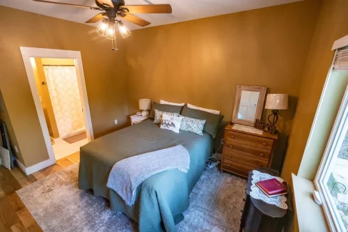 Warm-toned bedroom with golden walls, queen bed, wooden dresser, and ensuite bath visible through doorway.