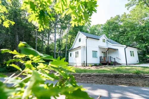 White modern farmhouse framed by lush green leaves and tall trees in the background.