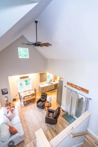 View from upstairs loft overlooking vaulted living room, dining area, and kitchen with ceiling fan and tall windows.