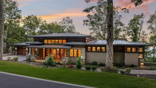 Contemporary mountain home with low rooflines, stone and wood siding, and glowing windows at dusk surrounded by trees.