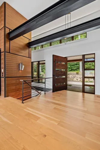 Modern entry with wood front door, steel beams, and open stair railing framed by natural light and warm wood accents.