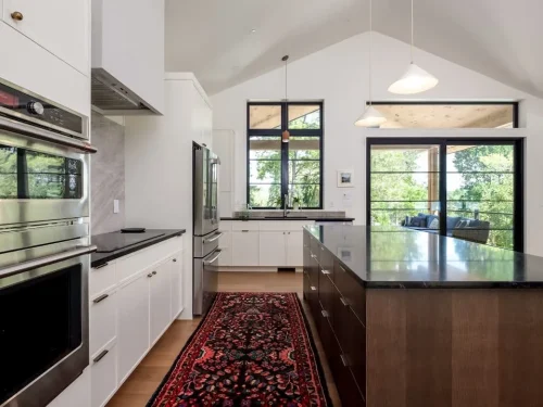 Bright kitchen with white cabinetry, dark countertops, a large island, vaulted ceiling, and black-framed windows overlooking the trees.