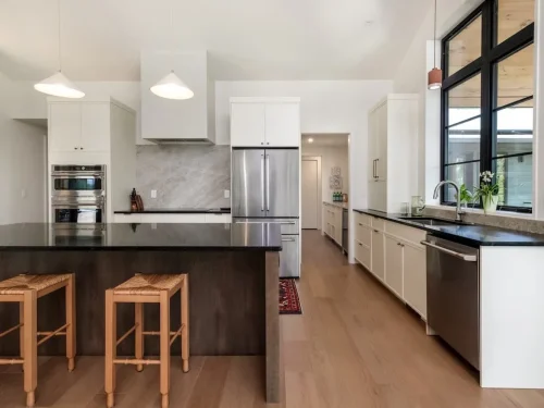 Modern kitchen with white cabinetry, dark island, stainless steel appliances, and large black-framed windows.
