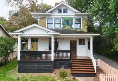 Front exterior of a renovated two-story white home with a covered porch, dark railings, wood steps, and colorful mural detail.
