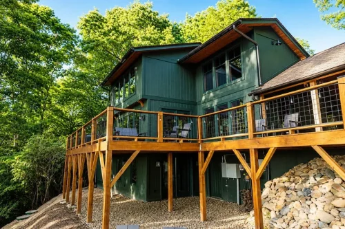 Rear exterior with dark green siding, large windows, and an elevated wood deck overlooking the wooded property.
