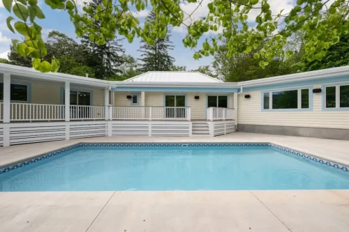 Renovated backyard pool with light exterior siding, white railings, covered deck, and concrete pool surround.