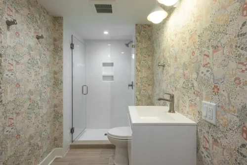 Renovated bathroom with patterned wallpaper, white vanity, and glass-enclosed walk-in shower.