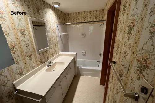 Before renovation bathroom with floral wallpaper, long vanity, and tub-shower combination.