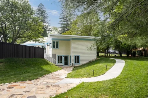 Renovated side exterior with light siding, blue trim, lower-level doors, and landscaped walkways.