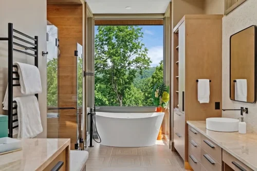 Modern bathroom with a freestanding tub positioned in front of a large window, wood cabinetry, a glass shower, and natural finishes.
