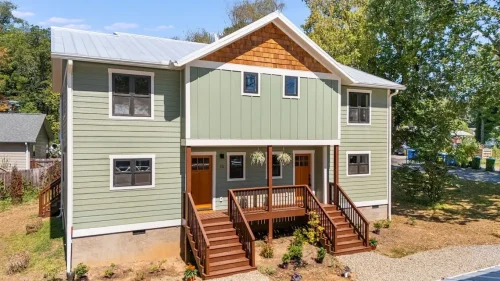 Two-story duplex with sage green siding, white trim, a metal roof, cedar shake accents, and covered wooden front porches.