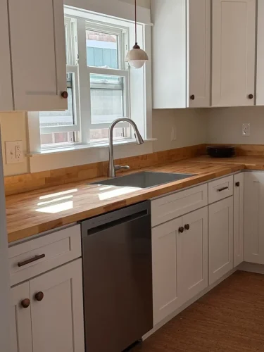 itchen with white shaker cabinets, butcher block countertops, a stainless steel dishwasher, and a sink beneath a window.