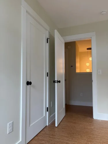 Interior hallway with light-colored walls, white paneled doors, black hardware, and natural-toned flooring.