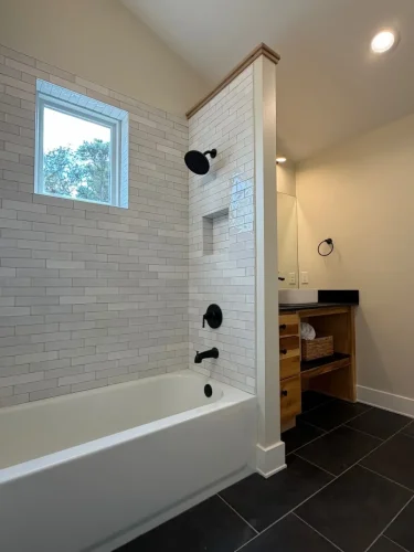 Bathroom with a tiled bathtub and shower, black fixtures, a window, dark floor tiles, and a natural wood vanity.