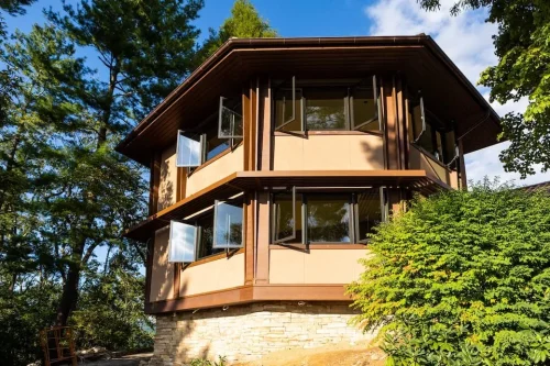 Multi-level home with warm wood detailing, stone foundation, broad roof overhangs, and rows of windows surrounded by trees.