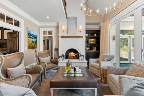 Elegant living room featuring a white brick fireplace, built-in shelving, upholstered seating, a gold chandelier, and large glass doors.