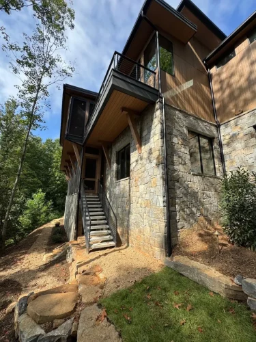 Stone and wood exterior with large windows, an upper balcony, exterior staircase, and wooded surroundings.