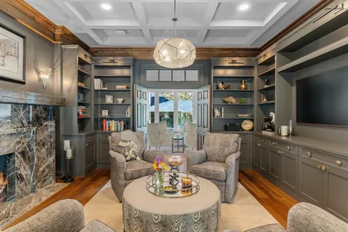 Library featuring dark gray built-in shelving, a marble fireplace, coffered ceiling, upholstered armchairs, and a geometric pendant light.