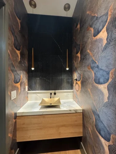Compact powder room with patterned wallpaper, a floating wood vanity, gold vessel sink, and dark tile accent wall.