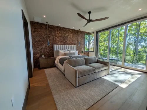 Bedroom with a wood accent wall, upholstered bed, sitting area, and large windows overlooking the wooded landscape.
