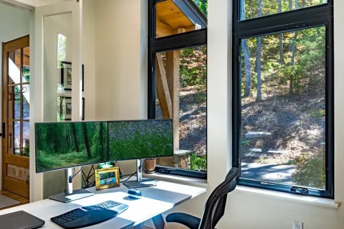 Home office with a desk facing large black-framed windows overlooking the wooded property.