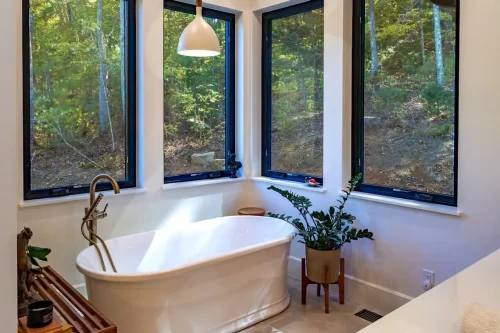 Bathroom with a freestanding soaking tub surrounded by large black-framed windows and wooded views.