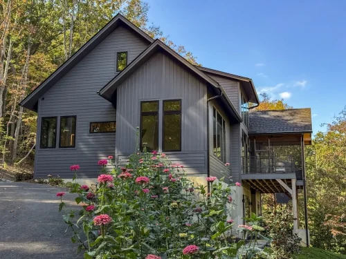 Side view of a dark gray mountain home with large black-framed windows, a raised deck, and wooded surroundings.