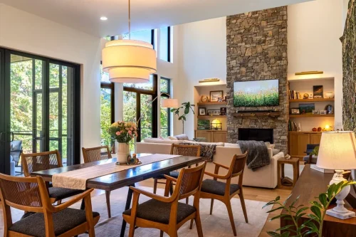 Open dining and living area with large windows, wood furnishings, and a floor-to-ceiling stone fireplace.