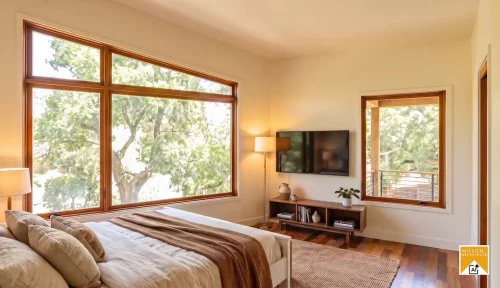 Bedroom with neutral bedding, large wood-trimmed windows overlooking trees, and a wall-mounted television above a low console.
