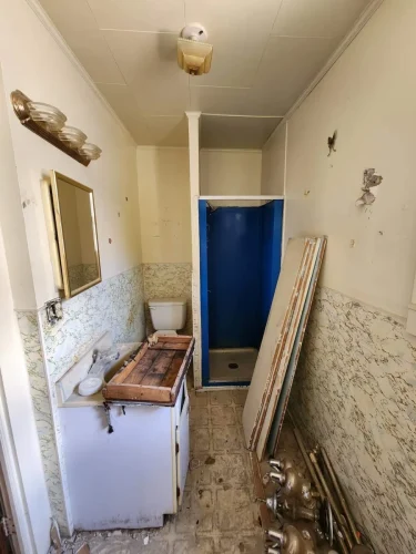 Old bathroom with damaged vanity, blue shower, and peeling walls.