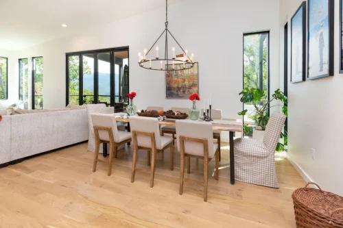 Modern dining room with wood floors, neutral upholstered chairs, and a round iron chandelier, opening to mountain views through large windows.