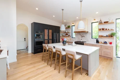 Bright modern kitchen with a marble island, pendant lighting, wood cabinetry, open shelving, and black built-in appliances.