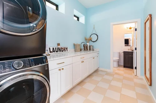 Laundry room with stacked washer and dryer, white cabinetry, farmhouse sink, and light blue walls with checkered tile flooring.