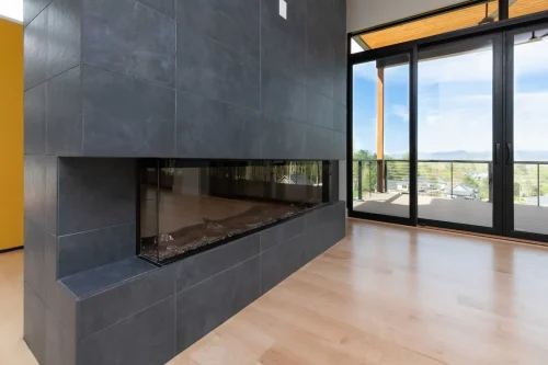 Contemporary double-sided fireplace with dark tile surround, light hardwood floors, and floor-to-ceiling windows opening to a mountain view deck.