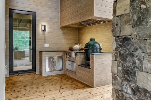Covered outdoor kitchen with built-in appliances, wood-look cabinetry, stone accents, and a ceramic grill.