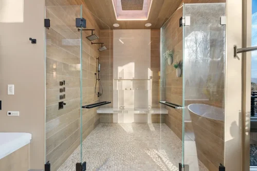 Large glass-enclosed walk-in shower with wood-look tile, pebble flooring, multiple showerheads, and a built-in bench.