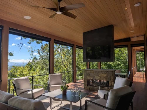 Outdoor living area with a wood ceiling, stone fireplace, mounted television, seating area, and wooded mountain views.